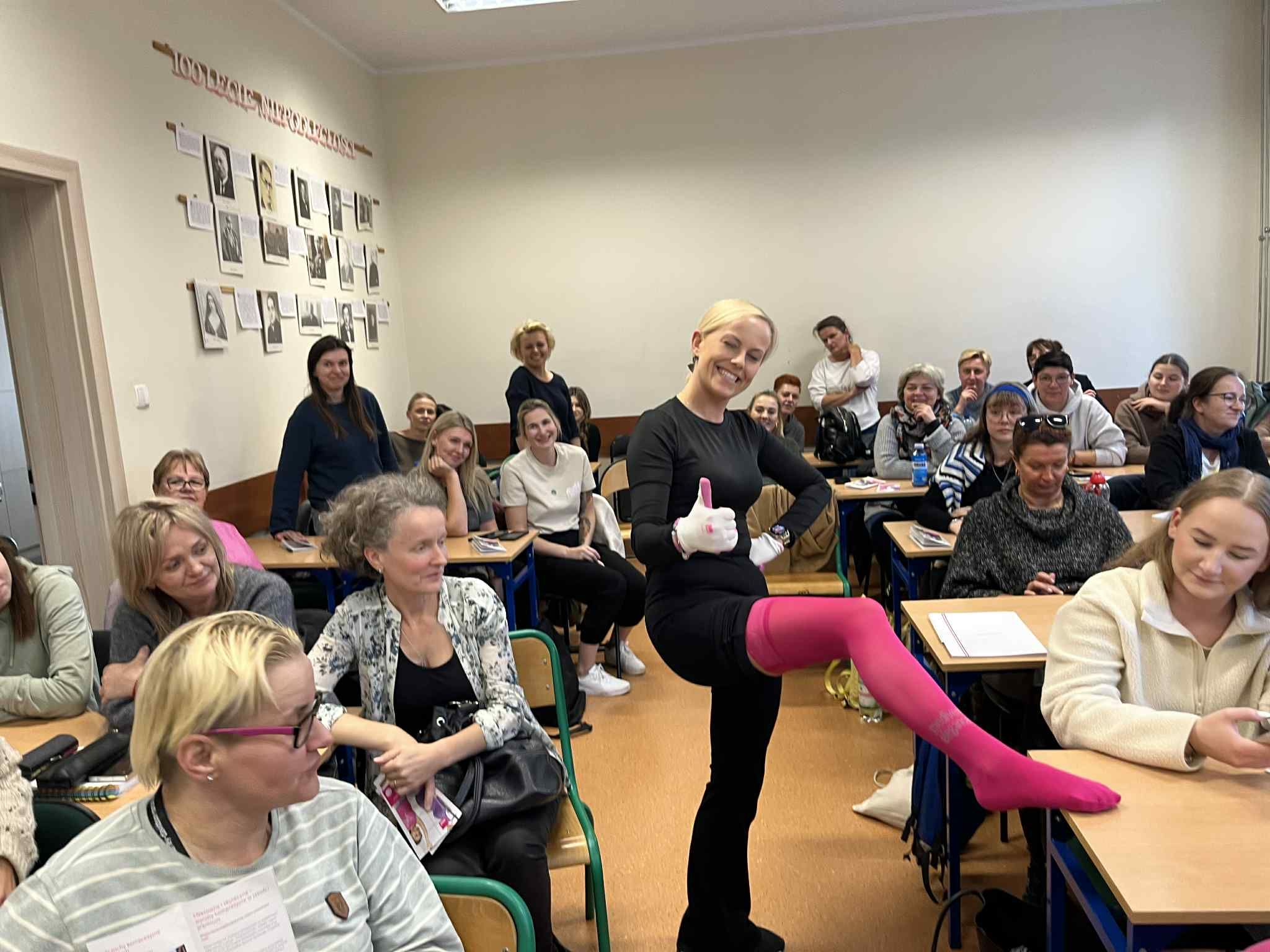 Zdjęcie w sali wykładowej z słuchaczką pozująca z różową pończochą. 