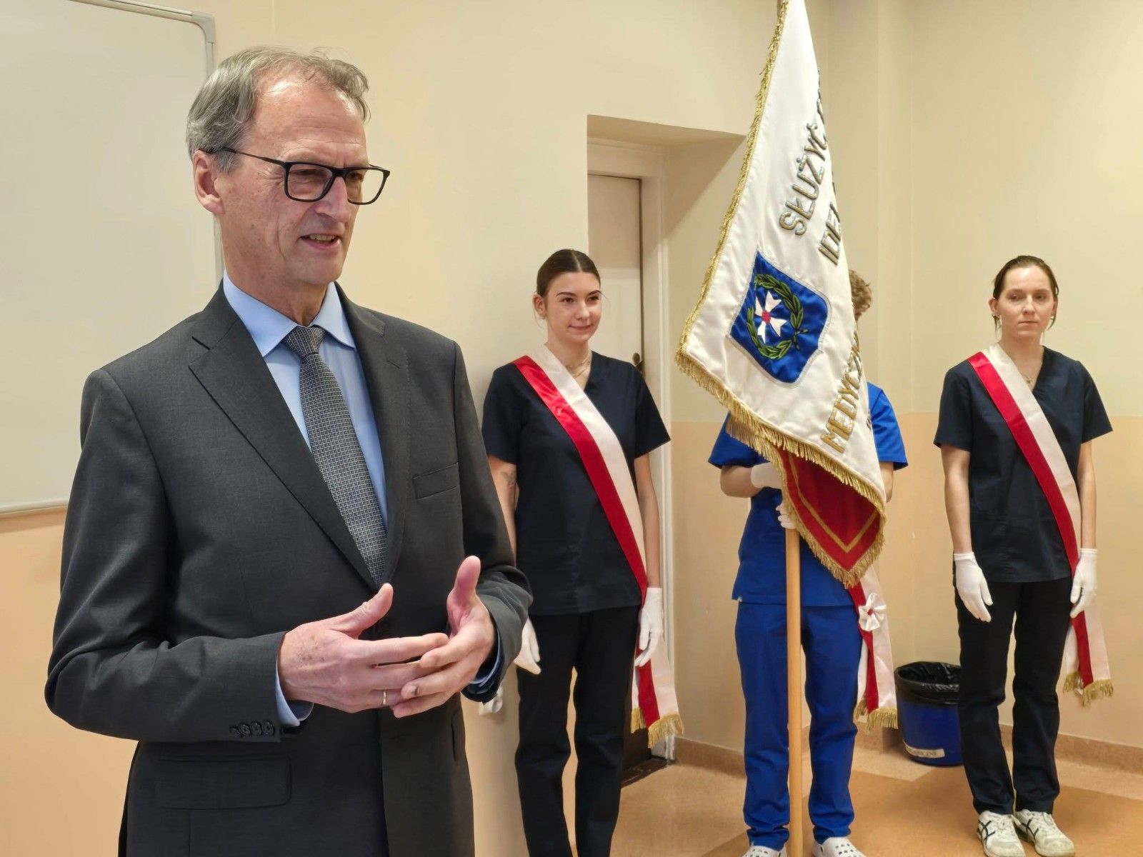 Widok na przemawiającego dyrektora szkoły wraz z pocztem sztandarowym w tle.