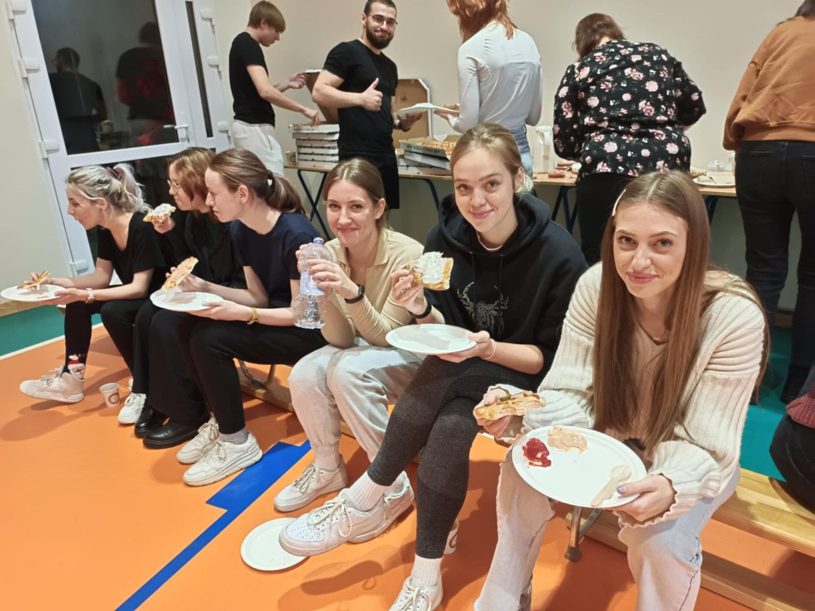 Widok na uczennice siedzące na ławce na hali i pożywające posiłek.