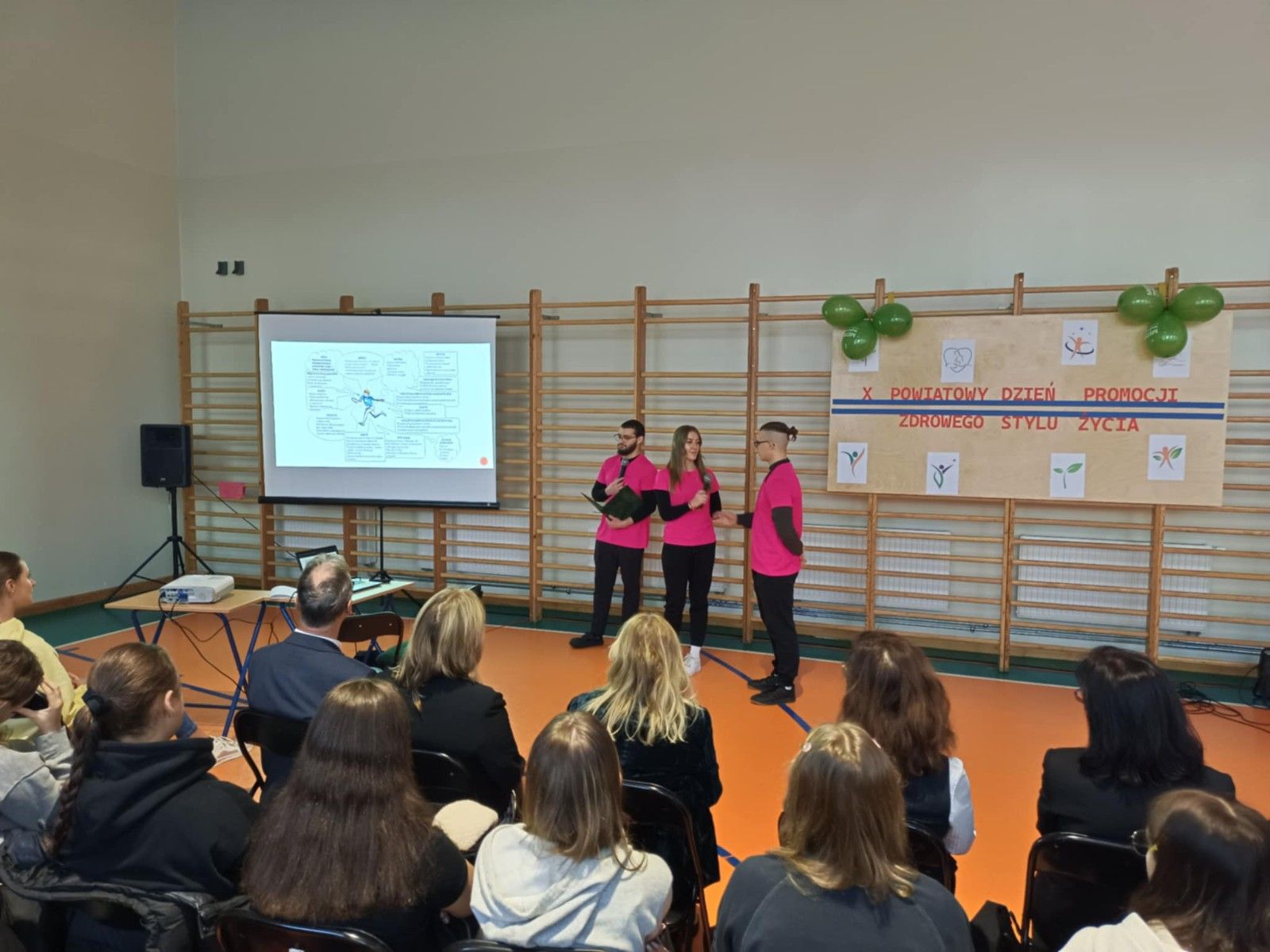 Uczniowie wraz z gośćmi prezentujący pokaz slajdów na hali sportowej. 