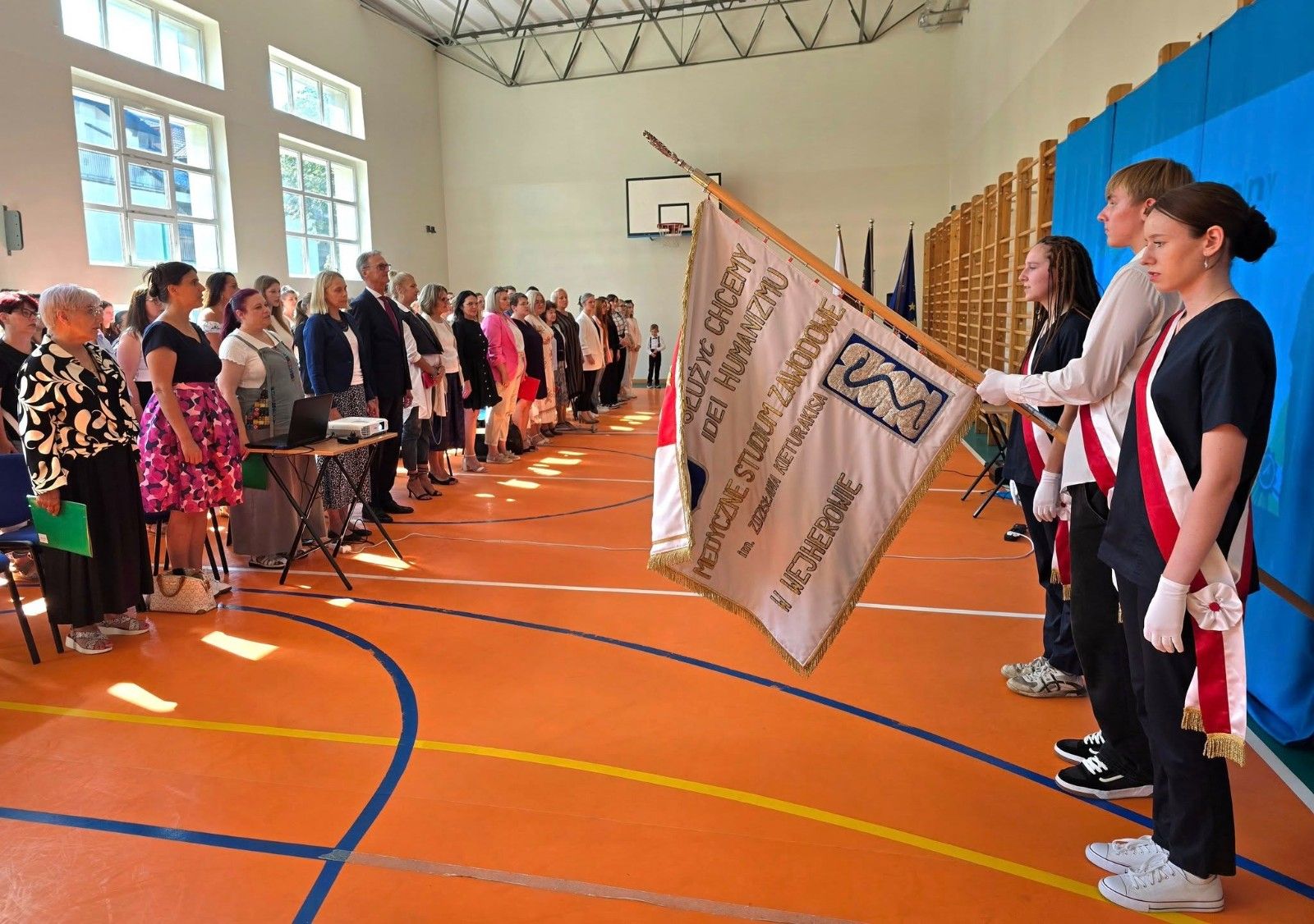 Widok na uczniów i nauczycieli na hali sportowej podczas śpiewu hymnu.