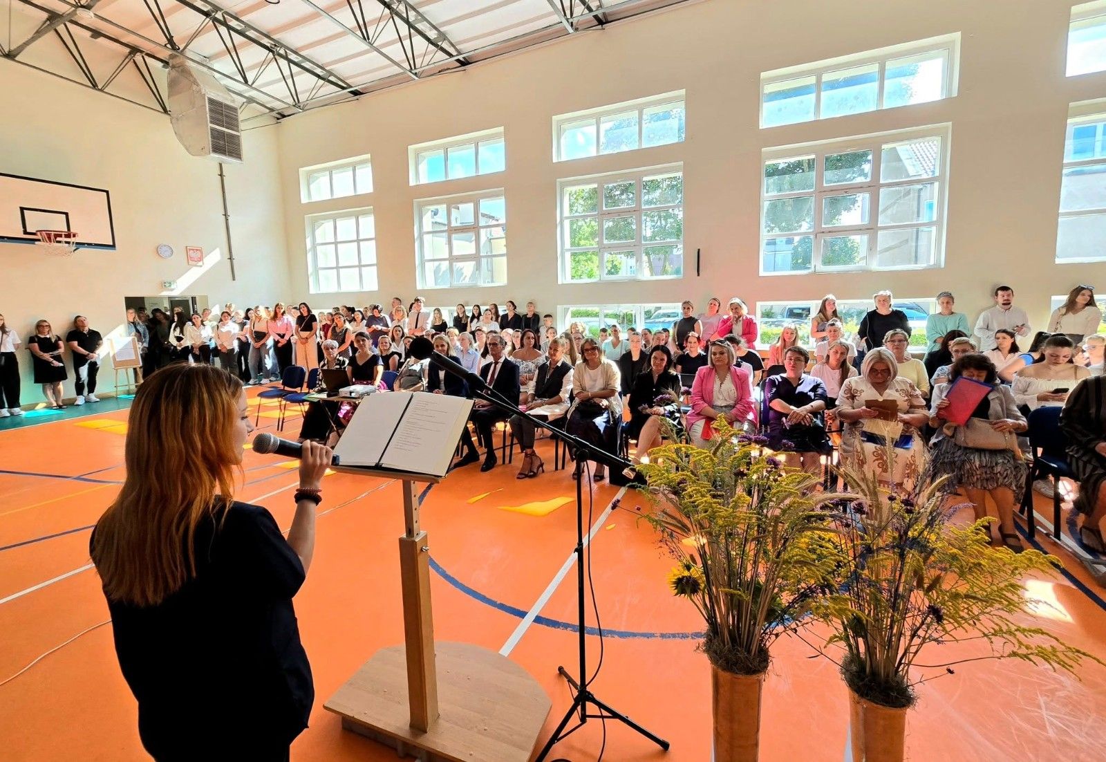 Widok na uczniów i nauczycieli na hali sportowej podczas przemówienia.